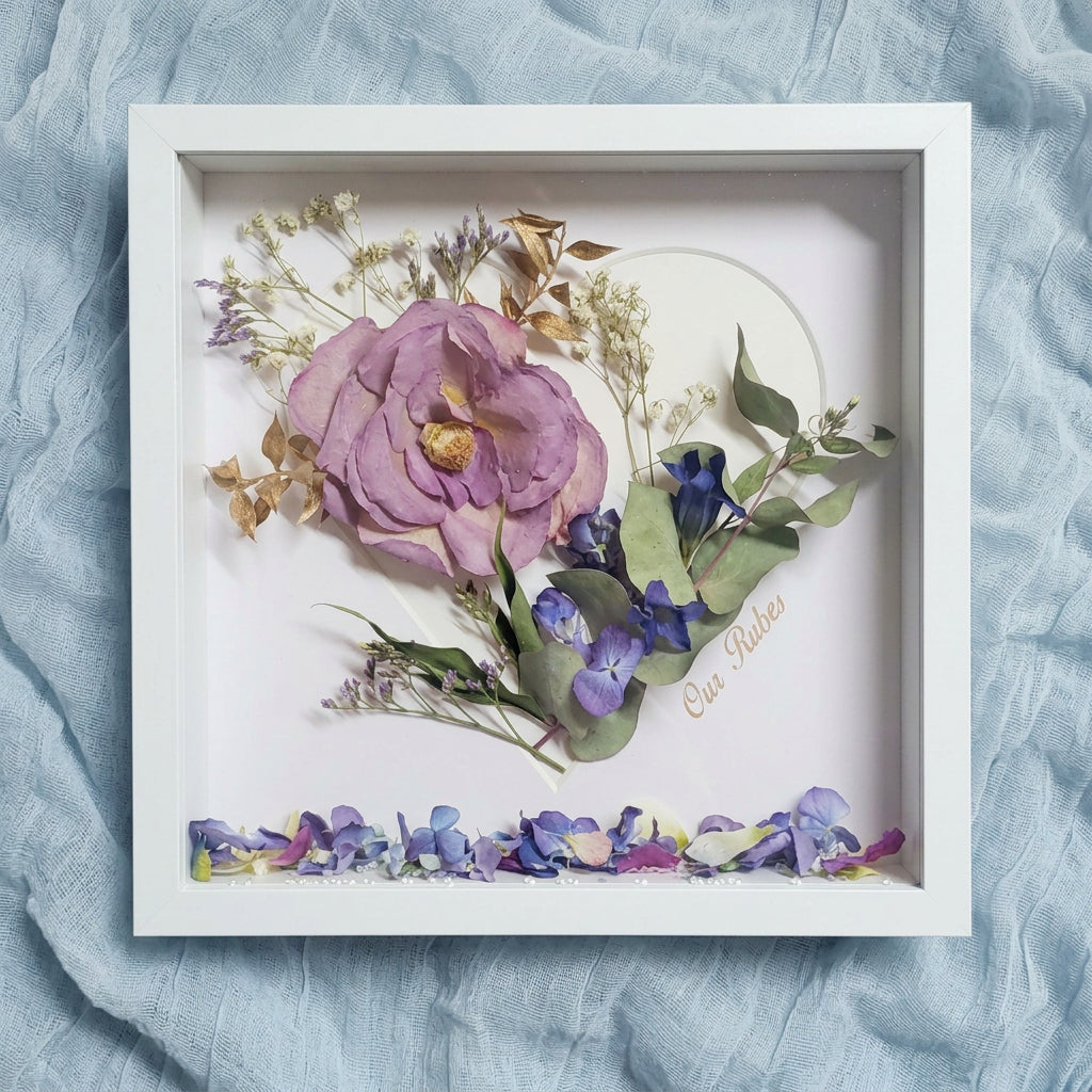 A Personalised square white shadow box frame containing a preserved floral arrangement with a large lavender flower, blue-purple blossoms, green leaves, and scattered flower petals along the bottom