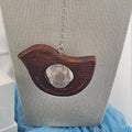 A wooden bird-shaped pendant necklace with a faceted clear crystal center, hanging on a silver chain from a beige fabric jewelry display bust.
