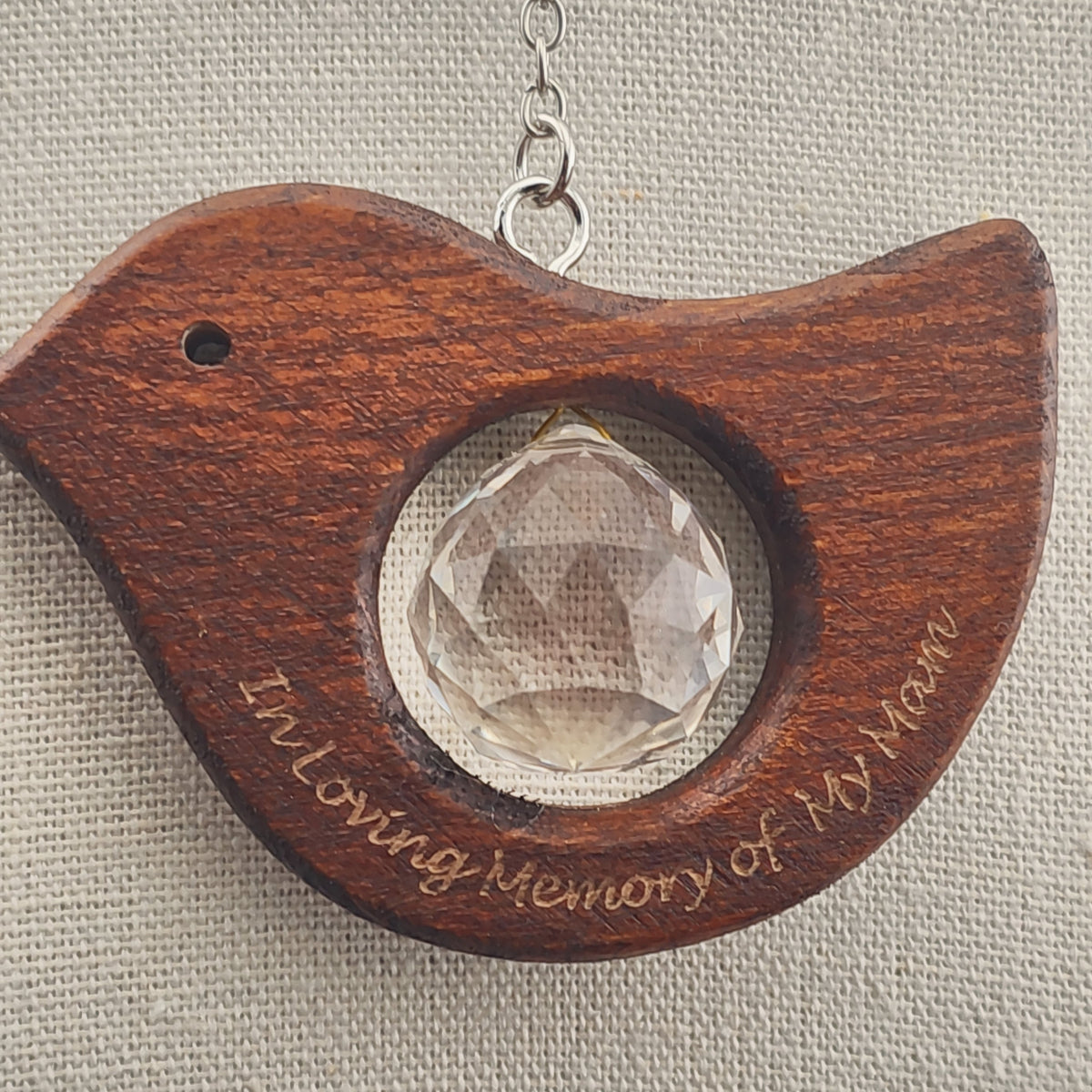 A wooden bird-shaped hanging ornament with a clear faceted crystal center and engraved text reading 'In Loving Memory of My Mom,' shown against a neutral textured fabric background.