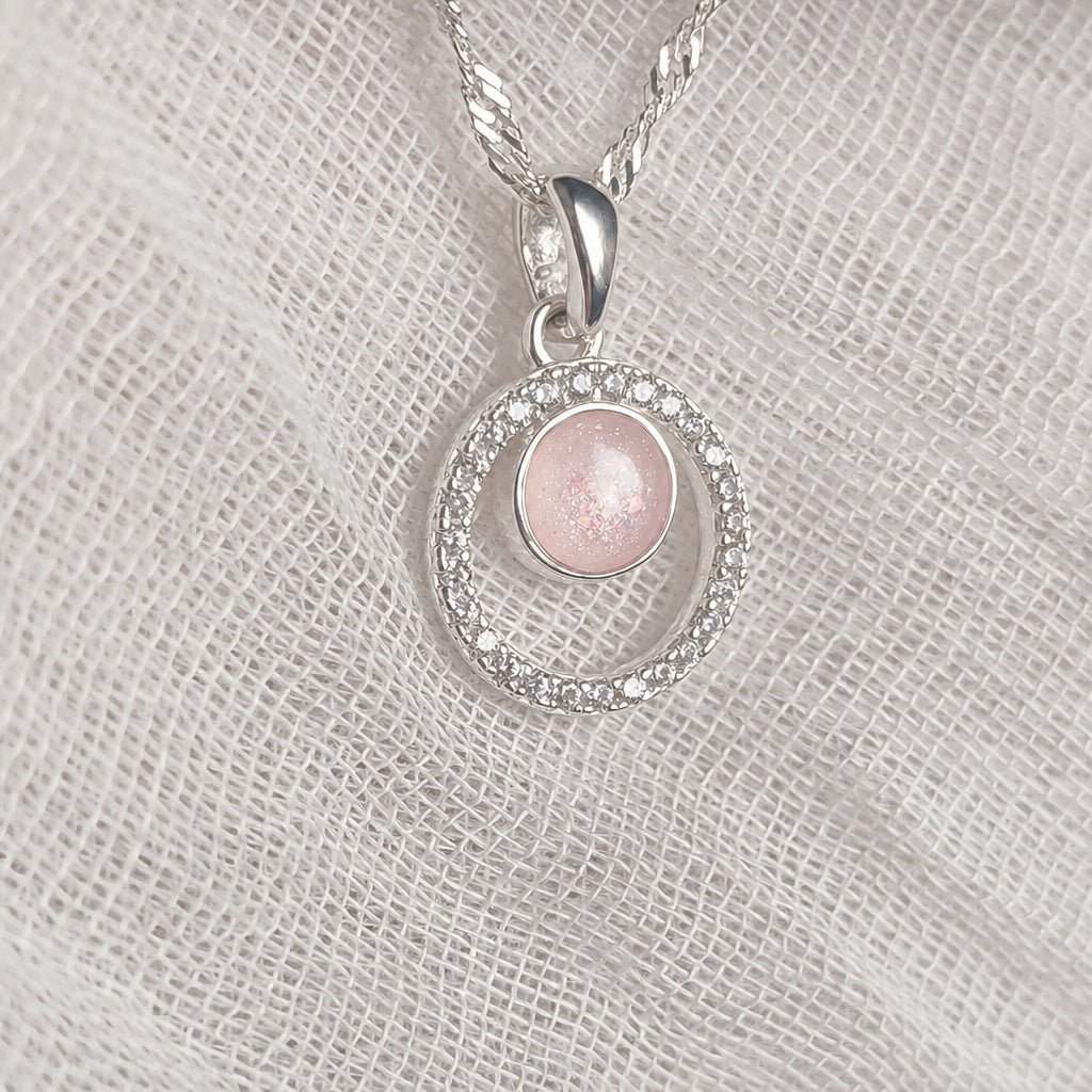 A silver pendant necklace featuring a ring design with a clear crystal halo and a central round iridescent white stone, displayed on a textured white fabric background.