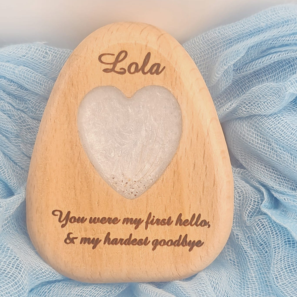 A personalized wooden keepsake plaque shaped like a teardrop, engraved with the name 'Lola' at the top and the quote 'You were my first hello, & my hardest goodbye' at the bottom. The center features a heart-shaped cutout containing a white textured substance.