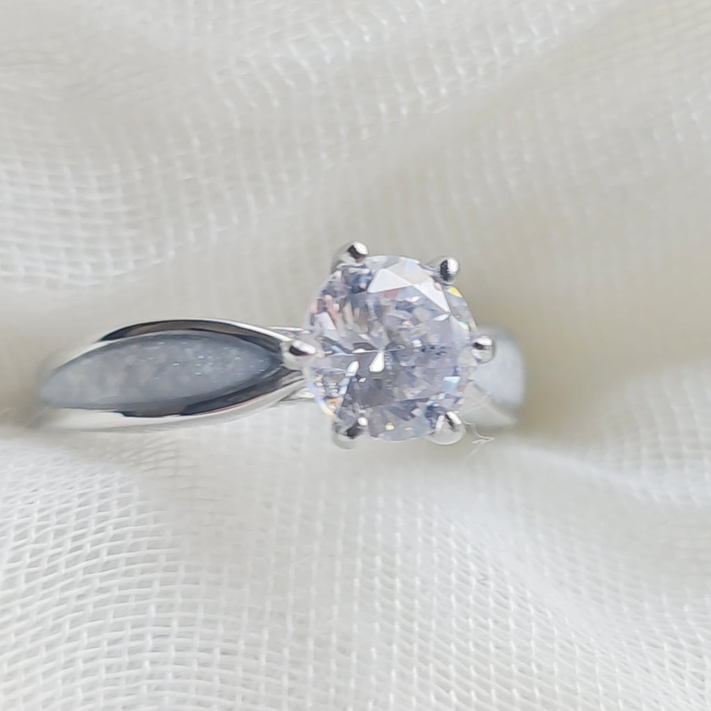 A silver solitaire ring with a round clear gemstone, resting on a textured white fabric background.