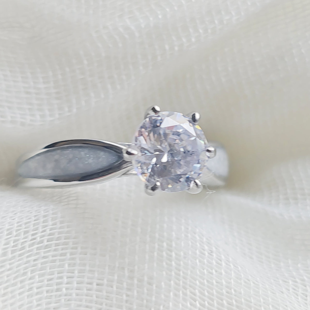 A silver solitaire ring with a round clear gemstone, resting on a textured white fabric background.