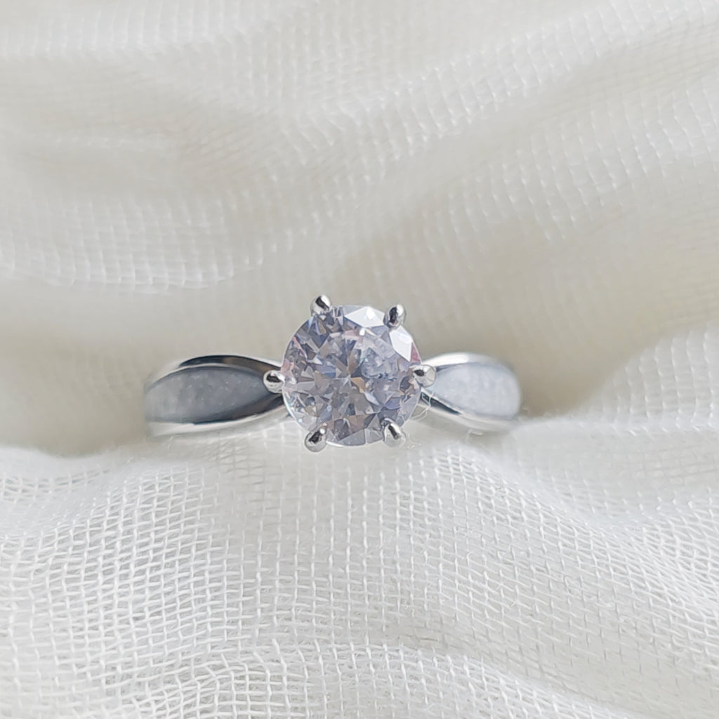 A silver solitaire engagement ring with a round clear gemstone, displayed on a textured white fabric background.