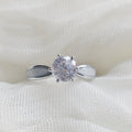 A silver solitaire engagement ring with a round clear gemstone, displayed on a textured white fabric background.