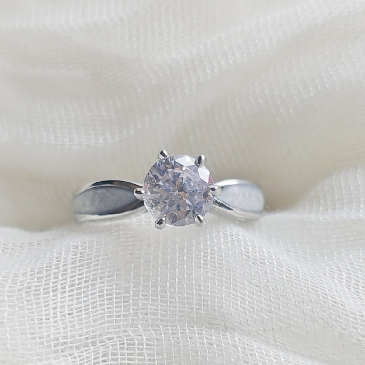 A silver solitaire engagement ring with a round clear gemstone, displayed on a textured white fabric background.
