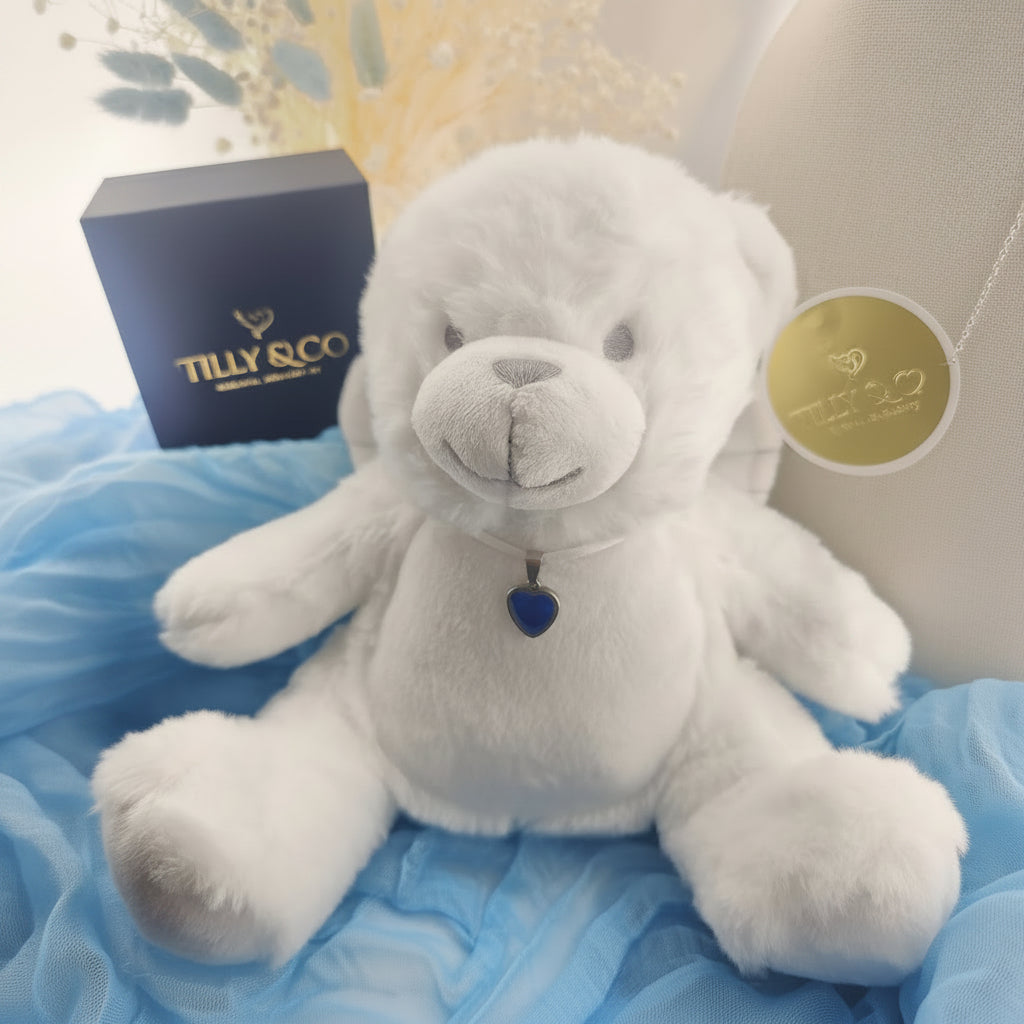 A white plush teddy bear with a blue heart-shaped pendant, displayed on a blue fabric surface with a dark blue Tilly & Co gift box and a round gold medallion in the background.