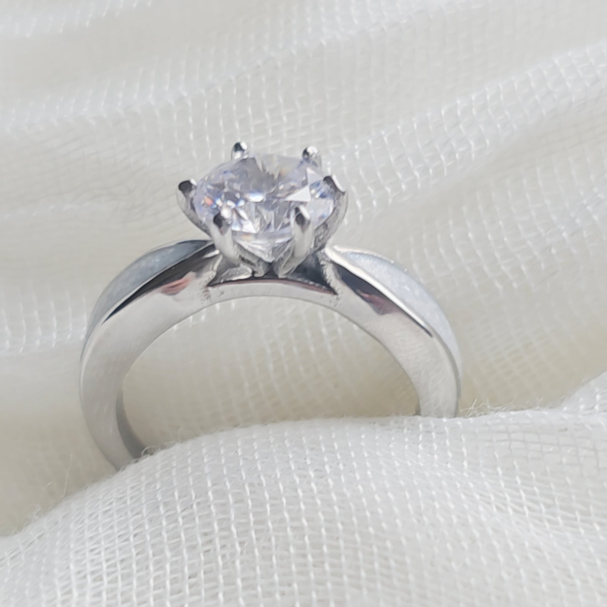 A silver solitaire engagement ring with a heart-shaped clear gemstone, resting on a textured white fabric background.