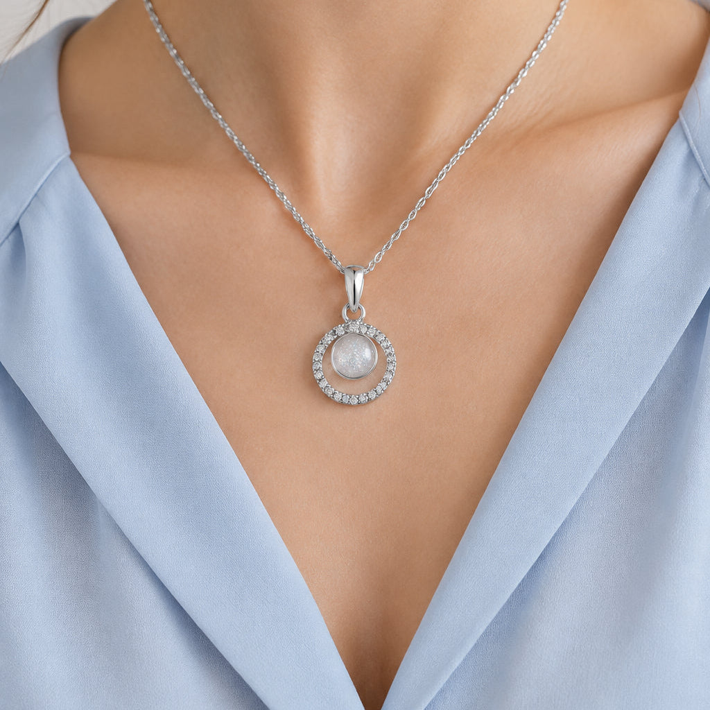 A silver necklace with a double-ring pendant, featuring a textured outer ring and a smooth, glittery inner circle, displayed on a white textured fabric background.