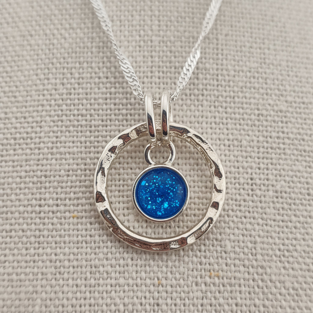 A silver necklace with a textured double-ring pendant and a small round iridescent charm, displayed on a beige woven fabric background.